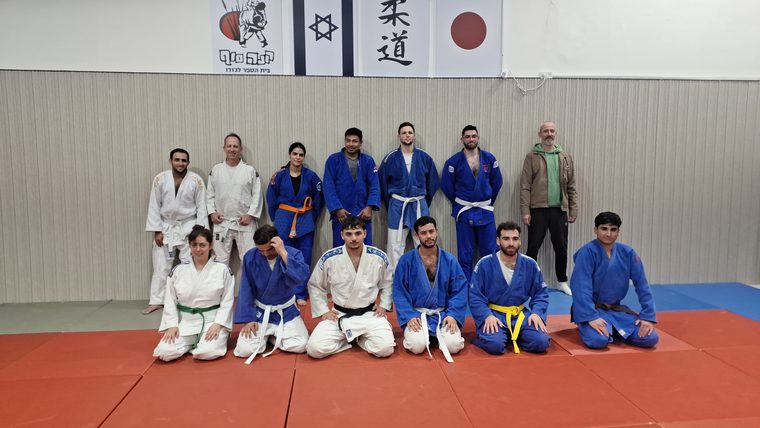 קבוצת ג׳ודופאן מצטלמת יחד מול שלט הג׳ודו של המועדון, מתאמנים בחגורות בצבעים שונים בשורה עומדת ושורה כורעת